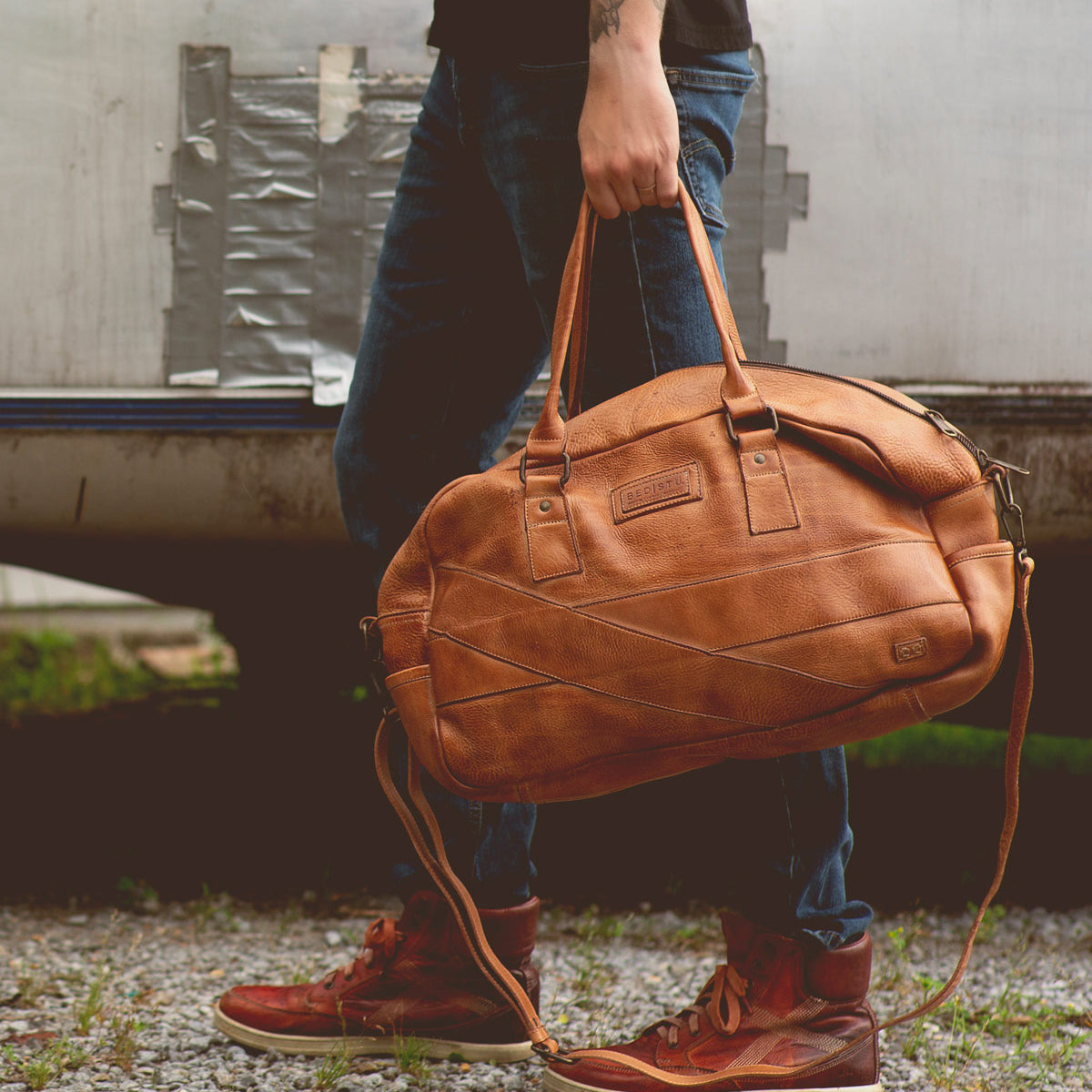 A person in jeans and red sneakers stands on gravel, holding the Bed|Stü "Luke" oversized duffle bag by its handles, crafted from rich vegetable tanned leather, against a backdrop of duct-taped metal.
