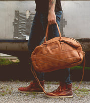 A person in jeans and red sneakers stands on gravel, holding the Bed|Stü "Luke" oversized duffle bag by its handles, crafted from rich vegetable tanned leather, against a backdrop of duct-taped metal.