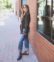 A woman wearing Bed|Stü's Lydyi black leather ankle boots with a cushioned insole, paired with a black leather jacket and blue cuffed jeans, stands on a brick sidewalk by a window, glancing over her shoulder.
