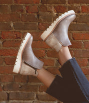 A person wearing the Bed|Stü Lydyi light brown leather ankle boots, featuring a zipper and thick white scalloped platform outsole, raises their legs against a red brick wall background; the boots are designed with a cushioned insole for added comfort.