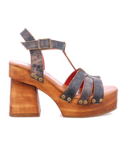 The Bed|Stü Lyxon is a single open-toe platform sandal featuring dark blue distressed straps, a chic t-strap design, metal rivet accents, an ankle buckle, and a chunky wooden heel.