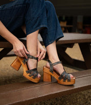 Person sitting on a picnic table bench, fastening the buckle on a pair of Bed|Stü Lyxon black and brown high-heeled sandals with a wooden platform heel and t-strap design, while wearing dark blue jeans.
