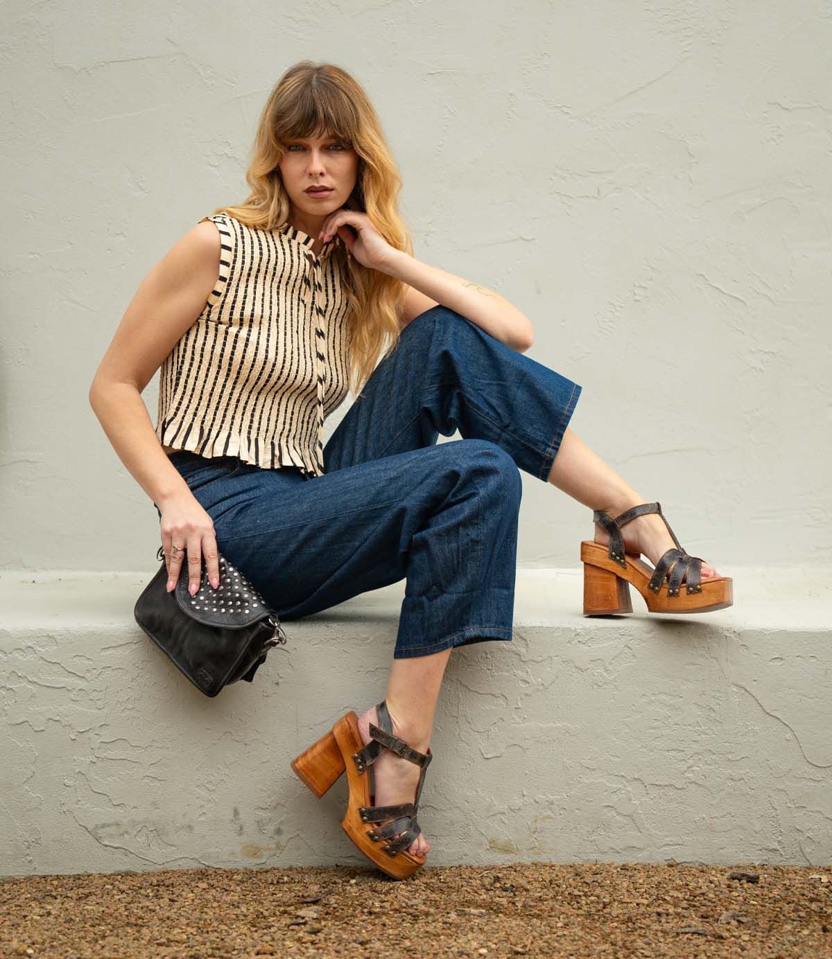 Woman with long hair poses seated on a ledge, wearing a striped sleeveless top, blue cropped pants, and Bed|Stü Lyxon wooden platform heel sandals with an open-toe t-strap design, while holding a black studded purse.