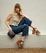 Woman with long hair poses seated on a ledge, wearing a striped sleeveless top, blue cropped pants, and Bed|Stü Lyxon wooden platform heel sandals with an open-toe t-strap design, while holding a black studded purse.