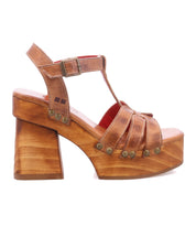 The Bed|Stü Lyxon is a brown leather platform sandal featuring a chunky wooden platform heel, metal rivet accents, and a buckle ankle strap. Styled in an open-toe silhouette, it is shown in a left-facing side profile against a white background.