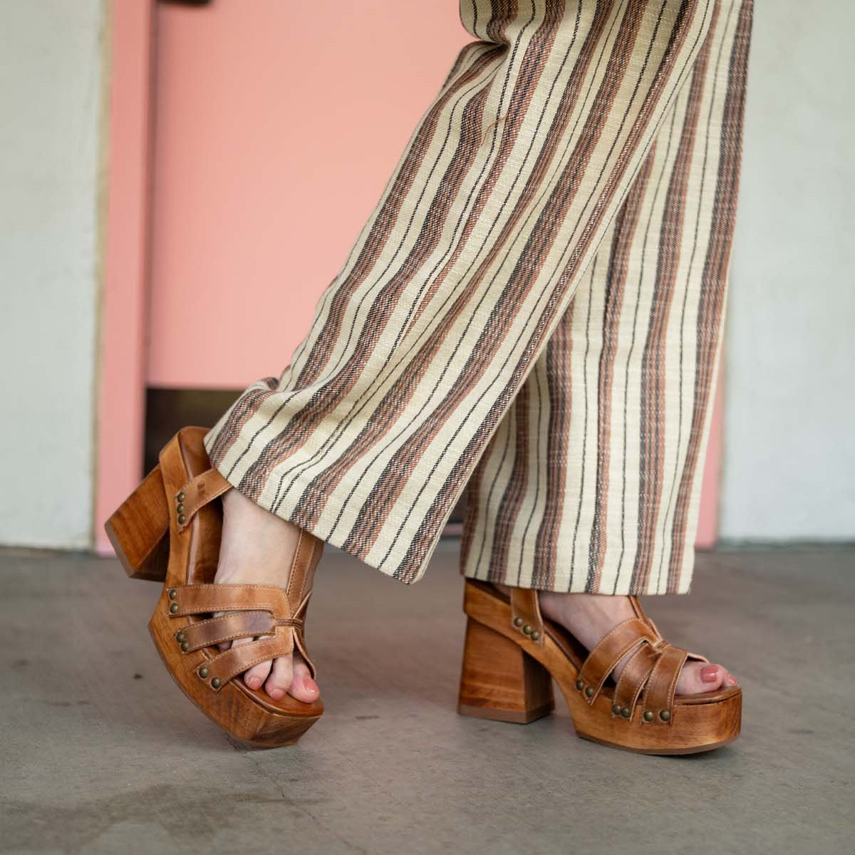 A person wearing the Bed|Stü Lyxon brown open-toe platform sandals with a t-strap design and beige striped pants, walking indoors on a gray floor with a pink door in the background.