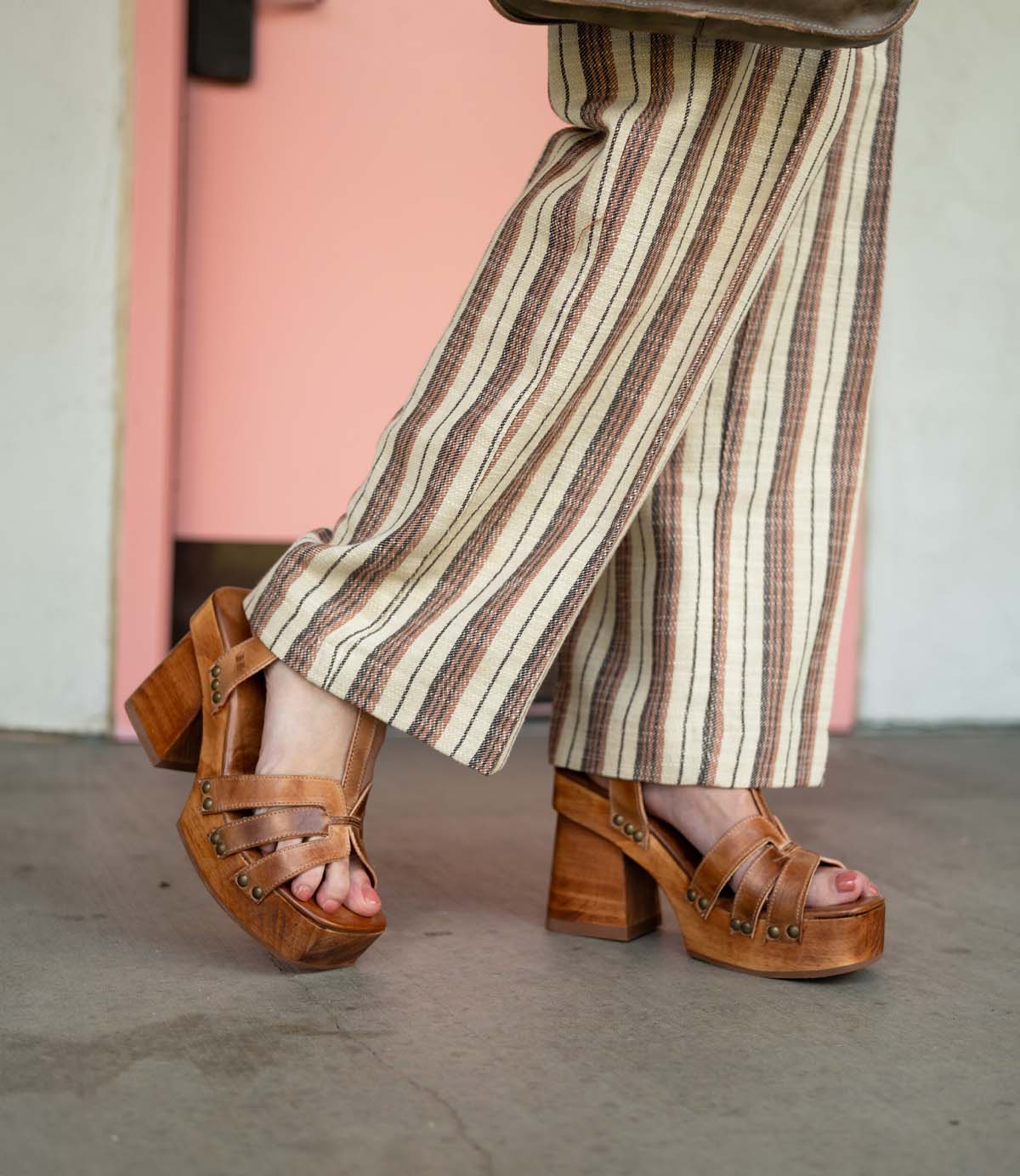 A person wearing the Bed|Stü Lyxon brown open-toe platform sandals with a t-strap design and beige striped pants, walking indoors on a gray floor with a pink door in the background.