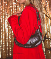 Wearing a striking red blazer, a person holds the Bed|Stü Madre shoulder bag—an edgy accessory detailed with metal studs—while standing before a shimmering gold sequin backdrop for an unforgettable, statement-making look.