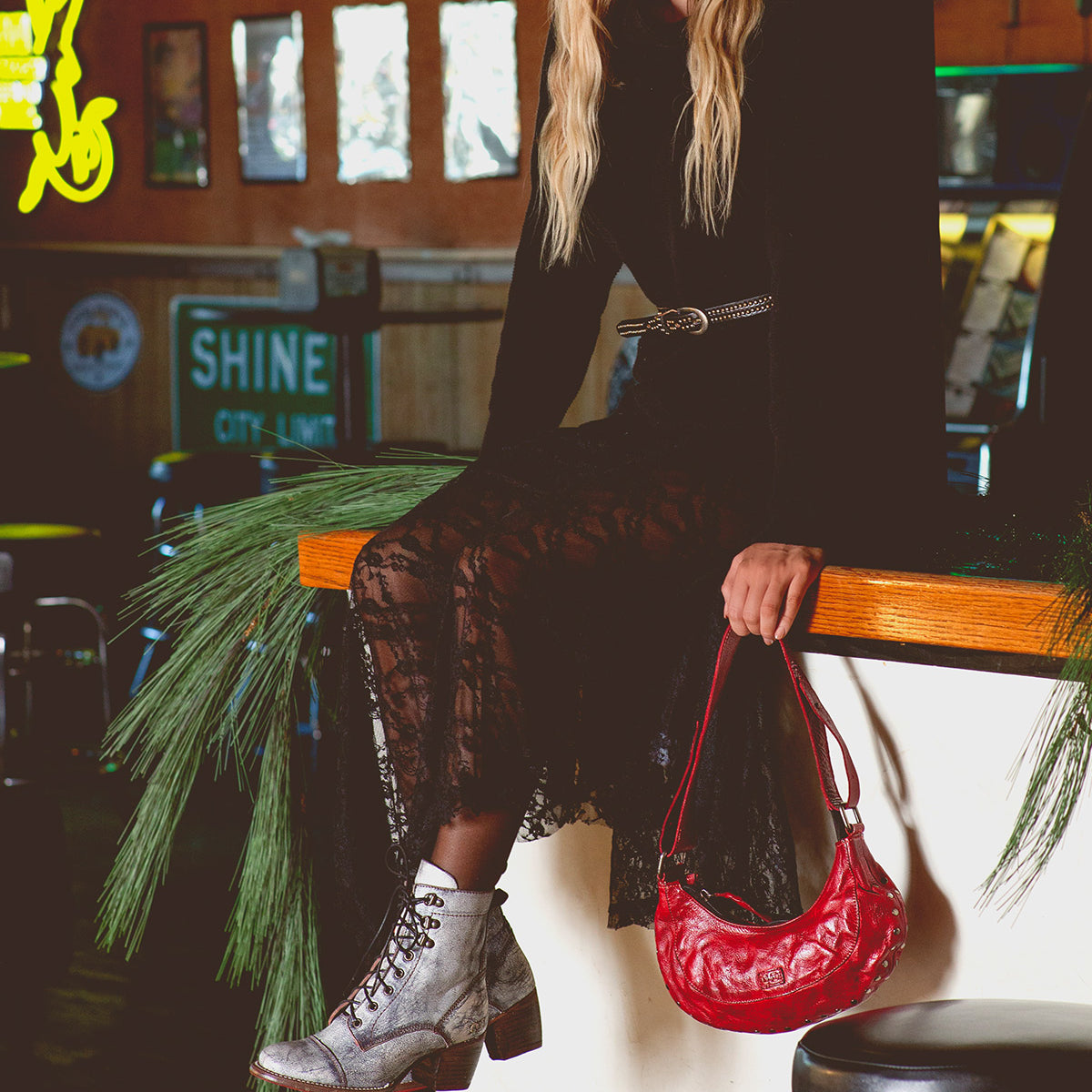 A woman—her face hidden—sits on a counter in a flowing black lace dress, holding the Bed|Stü Madre handbag, an eye-catching red piece embellished with metal studs. She pairs her look with gray lace-up boots, as pine branches and signs decorate the background.