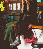 A woman—her face hidden—sits on a counter in a flowing black lace dress, holding the Bed|Stü Madre handbag, an eye-catching red piece embellished with metal studs. She pairs her look with gray lace-up boots, as pine branches and signs decorate the background.