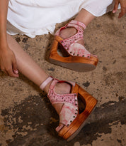 A person sits on a rough concrete floor in a white dress, wearing the Bed|Stü Magic pink tie-dye platform sandals featuring a peep-toe design, wooden soles, and studded trim.