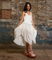 A woman in a white sleeveless dress and the Bed|Stü Magic platform sandals, featuring a handwoven leather upper, poses indoors against a weathered brick and plaster wall.