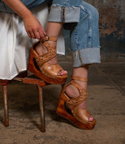 A person sits on a chair wearing rolled-up blue jeans and the Bed|Stü Magic platform wedge sandals, featuring a handwoven leather upper as they fasten the ankle strap. The sculpted wooden wedge and subtle peep-toe design of Magic add artisanal detail to their laid-back style.