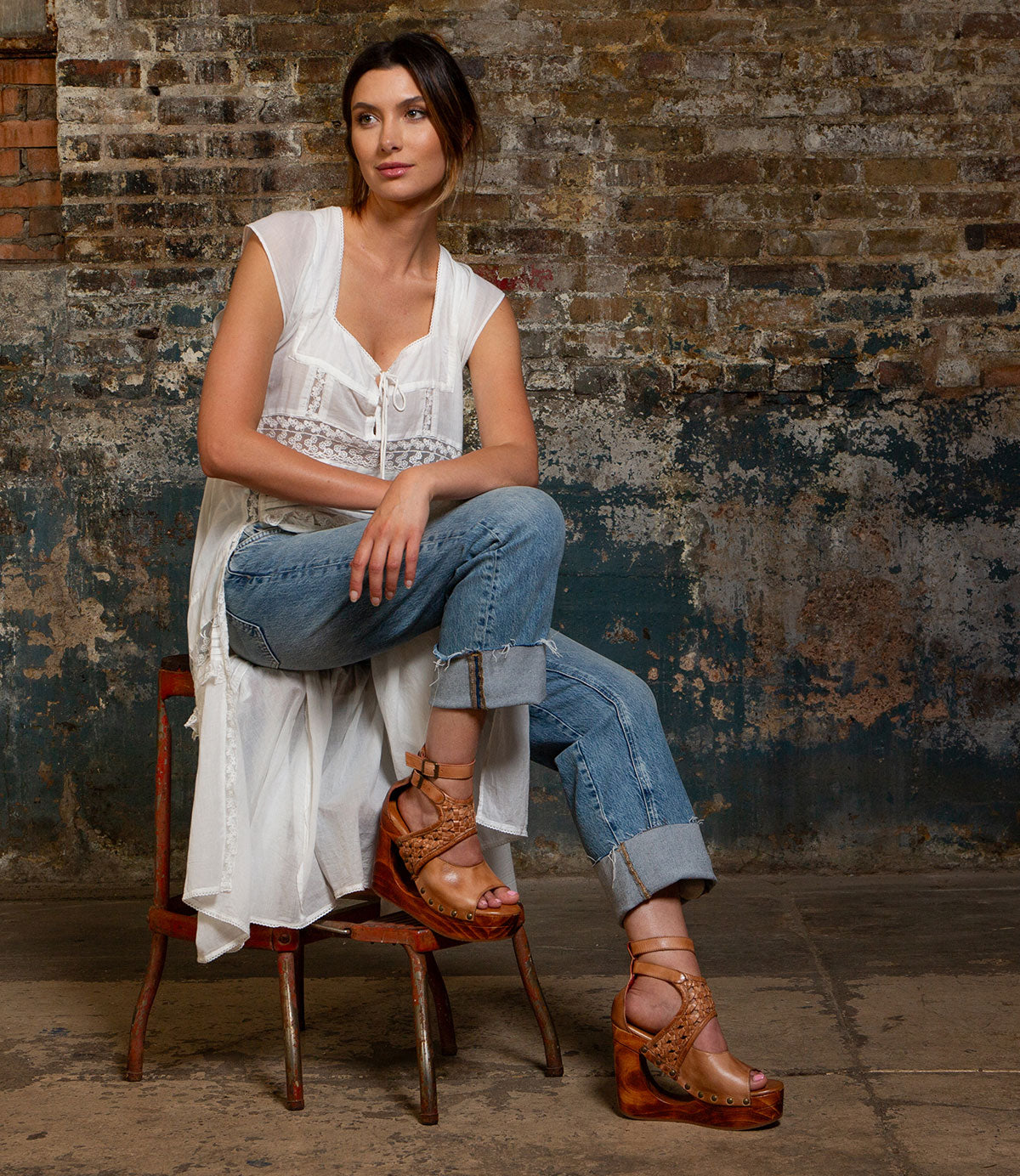 A woman sits on a chair against a textured brick wall, wearing a white sleeveless top, blue jeans, and Bed|Stü’s Magic sandals—brown platform shoes with a sculpted wooden wedge and handwoven leather upper.