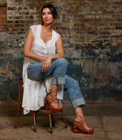 A woman sits on a chair against a textured brick wall, wearing a white sleeveless top, blue jeans, and Bed|Stü’s Magic sandals—brown platform shoes with a sculpted wooden wedge and handwoven leather upper.