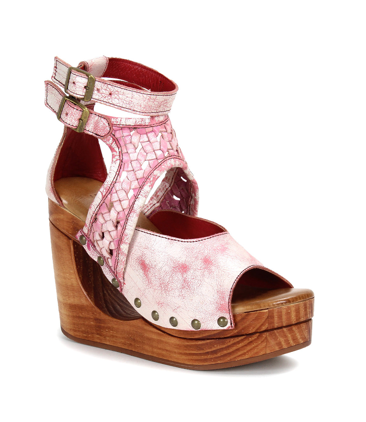 The Bed|Stü Magic is a pink and red leather wedge sandal showcasing a handwoven leather upper with woven straps, double ankle buckles, an open toe, and a sculpted wooden wedge platform sole detailed with metallic rivets.