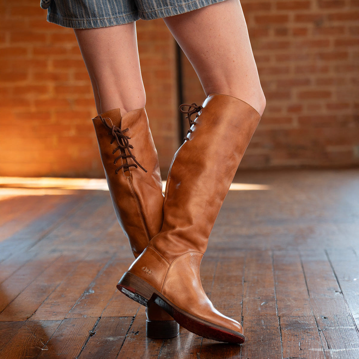 A person wearing the Bed|Stü Manchester knee-high boots in modern brown leather with a comfortable flat heel and striped shorts stands on a polished wooden floor with a brick wall in the background.