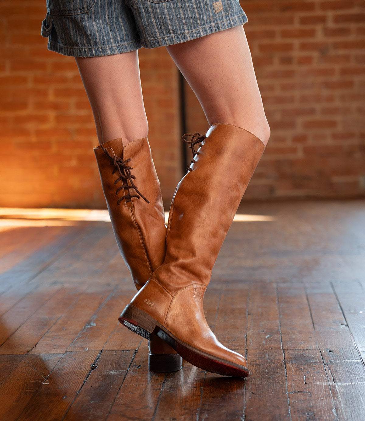 A person wearing the Bed|Stü Manchester knee-high boots in modern brown leather with a comfortable flat heel and striped shorts stands on a polished wooden floor with a brick wall in the background.
