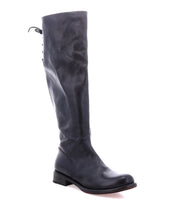 The Bed|Stü Manchester boot, crafted from sleek black leather, showcases a tall silhouette with an elegant back lacing detail and a comfortable flat heel. Presented against a white background, this modern classic effortlessly blends style and comfort for any occasion.