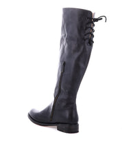 The Bed|Stü Manchester is a black knee-high leather boot featuring a low, comfortable flat heel, a side zipper, and lace-up detailing at the back of the calf. This modern classic boot is photographed against a white background.