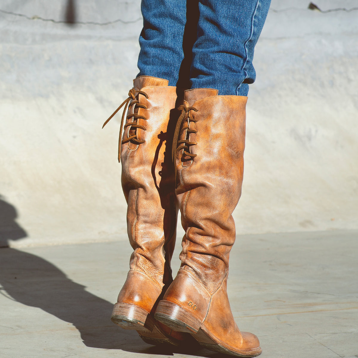 A person wearing blue jeans and the Bed|Stü Manchester boot—a modern classic, knee-high leather boot in distressed brown with back laces and a comfortable flat heel—stands on a concrete surface.