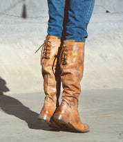 A person wearing blue jeans and the Bed|Stü Manchester boot—a modern classic, knee-high leather boot in distressed brown with back laces and a comfortable flat heel—stands on a concrete surface.