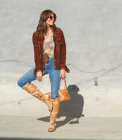 A woman wearing the Bed|Stü Manchester tan tall leather boots stands against a gray wall, styled in a brown jacket, lace top, blue jeans, sunglasses, and carrying a small brown handbag.