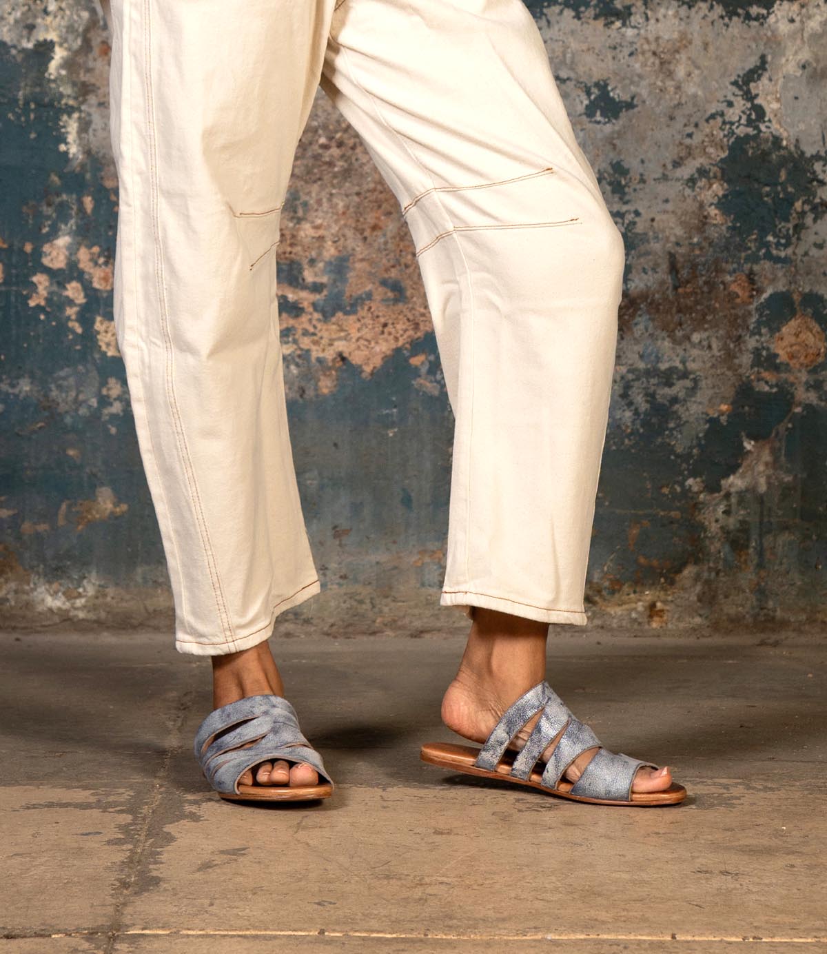 Wearing light-colored pants and the Bed|Stü Margin sandals—gray, strappy, and crafted with a leather upper—a person stands on a concrete floor against a worn, textured wall; an effortless addition to any summer wardrobe.