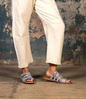 Wearing light-colored pants and the Bed|Stü Margin sandals—gray, strappy, and crafted with a leather upper—a person stands on a concrete floor against a worn, textured wall; an effortless addition to any summer wardrobe.