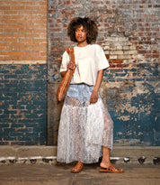Person standing by a textured brick wall, wearing a white t-shirt, sheer lace skirt over shorts, and Bed|Stü's Margin brown slide sandals with a leather upper—an effortless look for any summer wardrobe—while holding a brown shoulder bag.