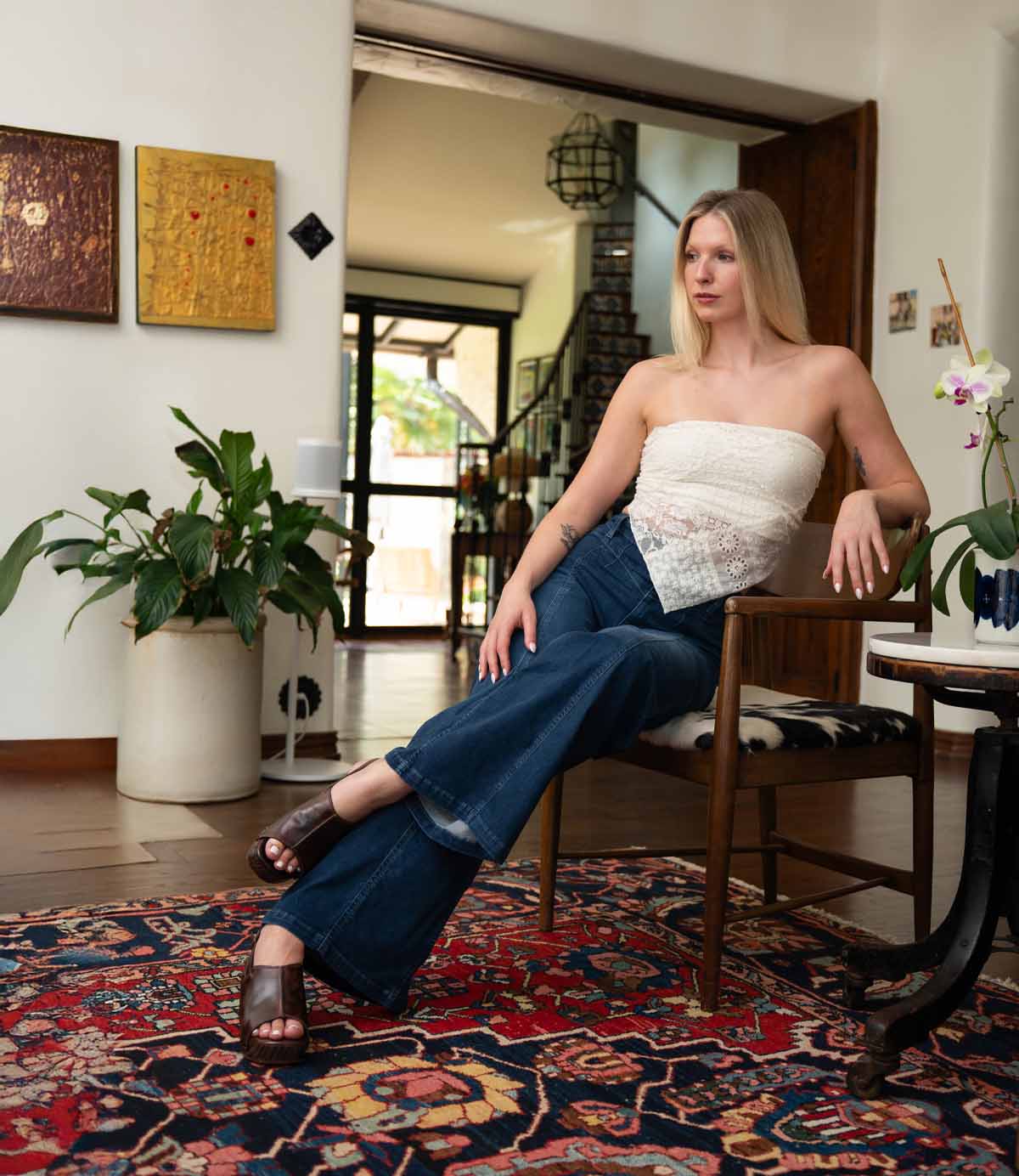 A woman in a white strapless top and blue jeans sits on a chair in a living room adorned with plants, artwork, and a patterned rug, her feet comfortably slipped into the Marie heeled clogs by Bed|Stü, crafted from vegetable tanned leather.