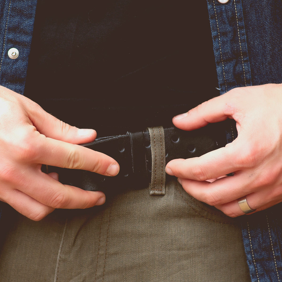 Wearing green pants paired with a denim jacket, a person fastens the Bed|Stü Mccoy—a black leather strap featuring a durable metal buckle—and sports a ring on their left hand.
