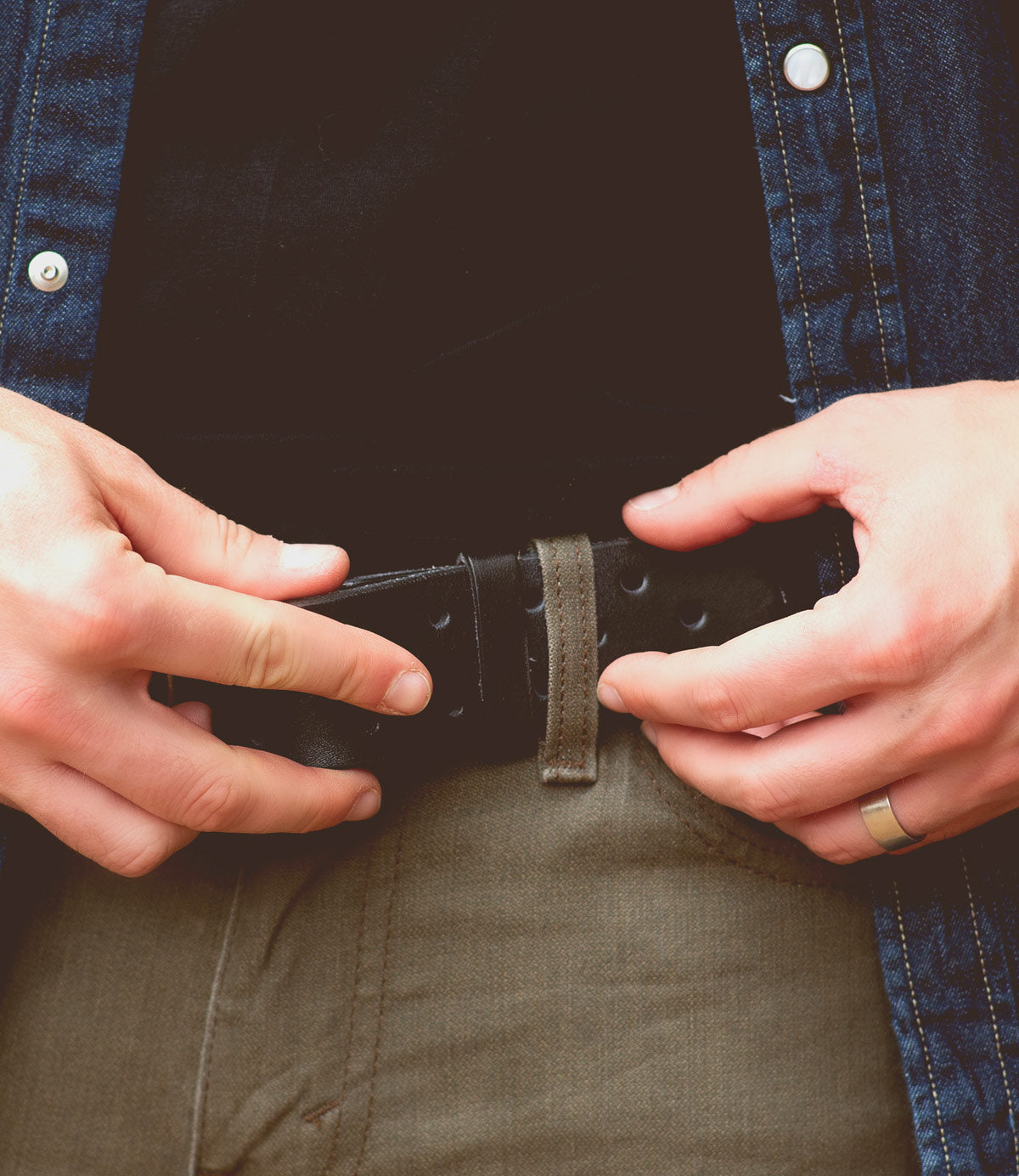 Wearing green pants paired with a denim jacket, a person fastens the Bed|Stü Mccoy—a black leather strap featuring a durable metal buckle—and sports a ring on their left hand.