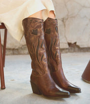 A person wearing cream pants and the Bed|Stü Meadow brown leather cowgirl boots with black decorative stitching sits on a chair on a concrete floor.
