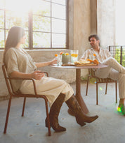 Two people sit at a small round table in a sunlit room, enjoying drinks and pastries near large windows. One of them casually rests her feet on the chair’s rung, showcasing her stylish Bed|Stü Meadow leather cowgirl boots that catch the morning light.