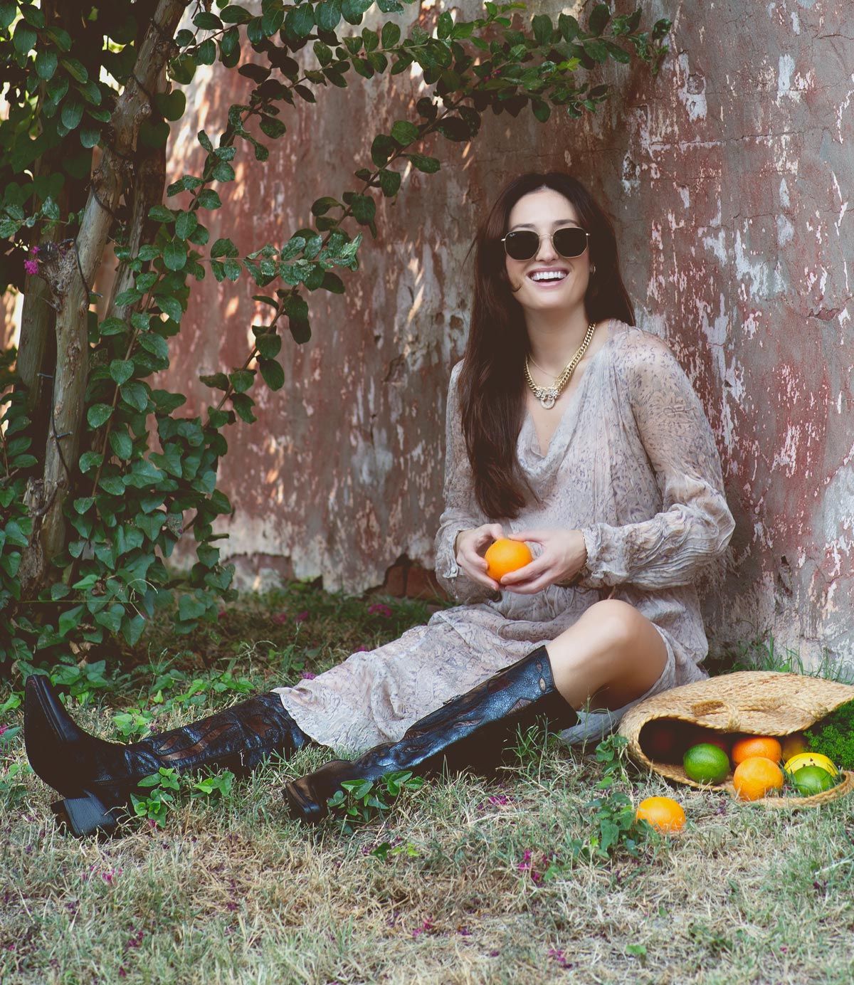 A woman wearing sunglasses and Bed|Stü’s handcrafted Italian "Meadow" boots sits on the grass by a wall, holding an orange beside a woven basket of spilled fruit.