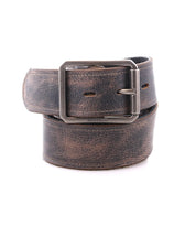 The Bed|Stü Meander is a rolled-up, distressed leather belt featuring an antique metal buckle and a rugged look, displayed on a white background.