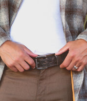 A person wearing a white shirt, plaid jacket, and brown pants is fastening the rugged Meander belt by Bed|Stü. Their hands are visible, with a metallic ring on one finger.