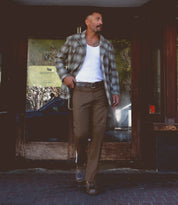 A man wearing a plaid jacket, white tank top, brown pants secured with the Bed|Stü Meander rugged belt featuring an antique metal buckle, and brown shoes walks on a sidewalk in front of a glass door.
