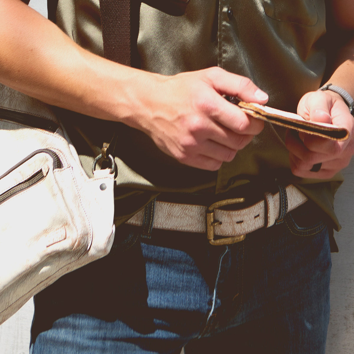 A person wearing a green shirt and blue jeans holds a phone and a wallet, with the Bed|Stü Meander beige shoulder bag hanging across their body. A rugged belt with an antique metal buckle completes the look, adding a touch of character to the casual outfit.