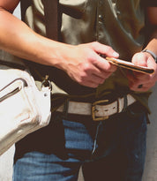 A person wearing a green shirt and blue jeans holds a phone and a wallet, with the Bed|Stü Meander beige shoulder bag hanging across their body. A rugged belt with an antique metal buckle completes the look, adding a touch of character to the casual outfit.