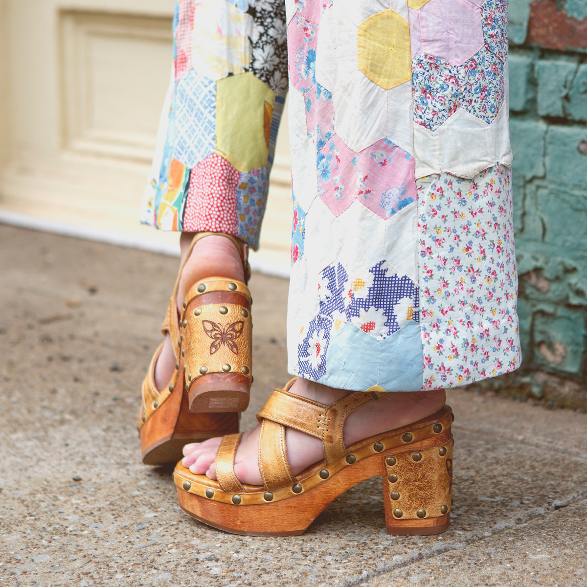 A person wearing patchwork pants and Bed|Stü's Mediation brown wooden platform sandals with leather straps and stud details stands on a sidewalk near a brick wall and cream-colored door.