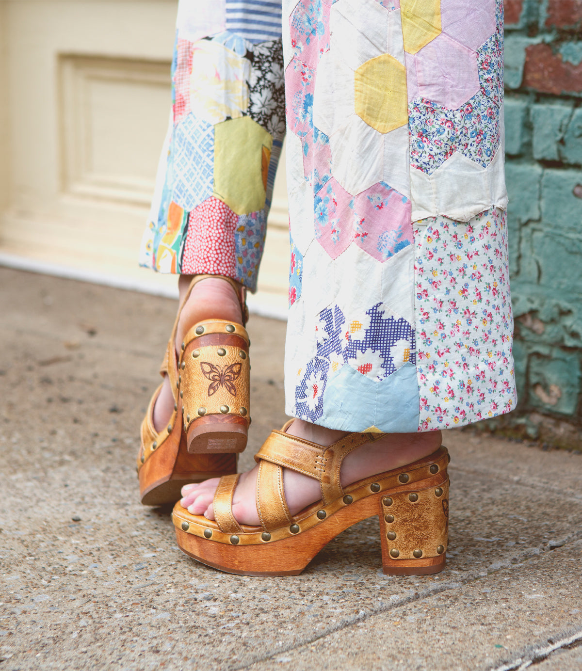 A person wearing patchwork pants and Bed|Stü's Mediation brown wooden platform sandals with leather straps and stud details stands on a sidewalk near a brick wall and cream-colored door.