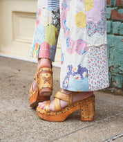 A person wearing patchwork pants and Bed|Stü's Mediation brown wooden platform sandals with leather straps and stud details stands on a sidewalk near a brick wall and cream-colored door.