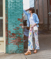 Leaning against a weathered brick wall on the sidewalk, a person in sunglasses and patterned pants completes their look with a blue shirt, white top, crossbody bag, and Mediation heeled slingback sandals with cushioned insoles by Bed|Stü.