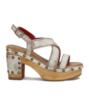 The Mediation by Bed|Stü is a chunky-heeled slingback sandal featuring wide metallic leather straps, a wooden sole accented with brass studs, a cushioned insole for extra comfort, and an adjustable ankle buckle.