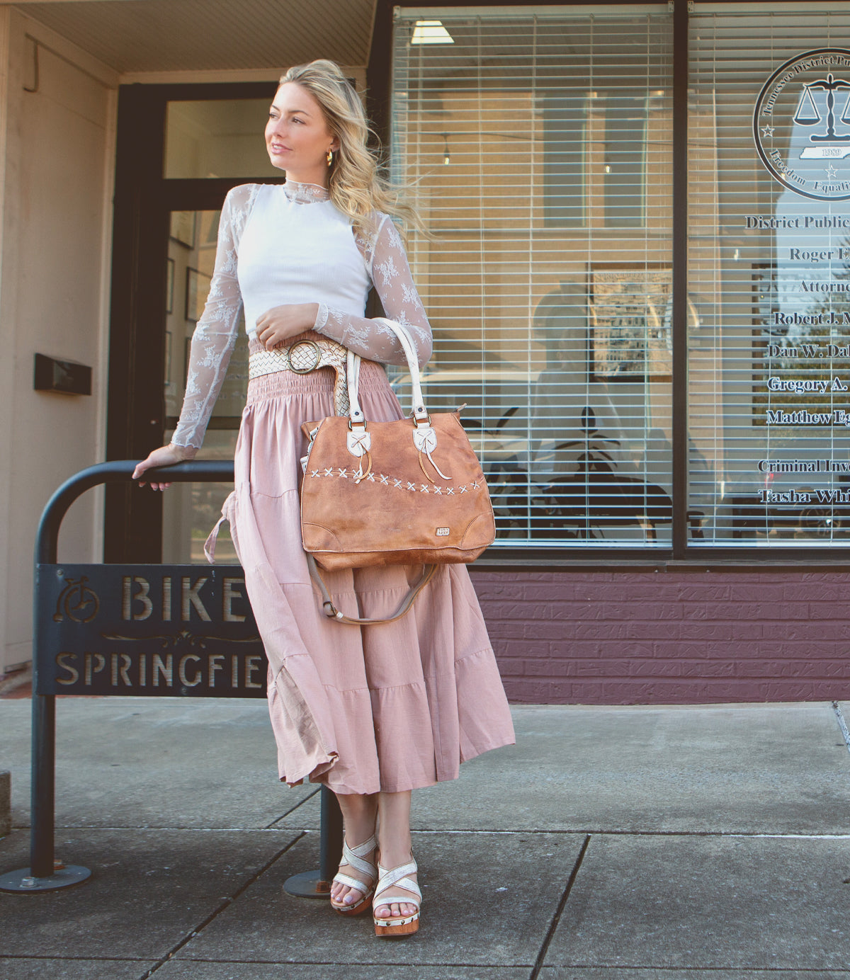 A woman in a white long-sleeve top and pink skirt stands on the sidewalk with a law office window behind her, carrying a brown handbag. She completes her look with Bed|Stü's Mediation heeled slingback sandals, which feature elegant leather straps and a cushioned insole for all-day comfort.