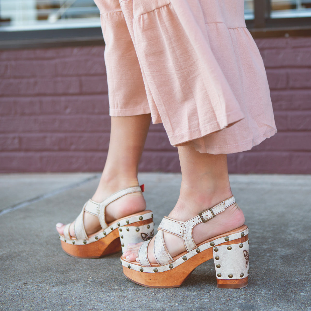 Wearing the Bed|Stü Mediation beige heeled slingback sandals with cushioned insoles and wooden soles, a person in light pink wide-leg pants stands on a sidewalk near a brick wall.