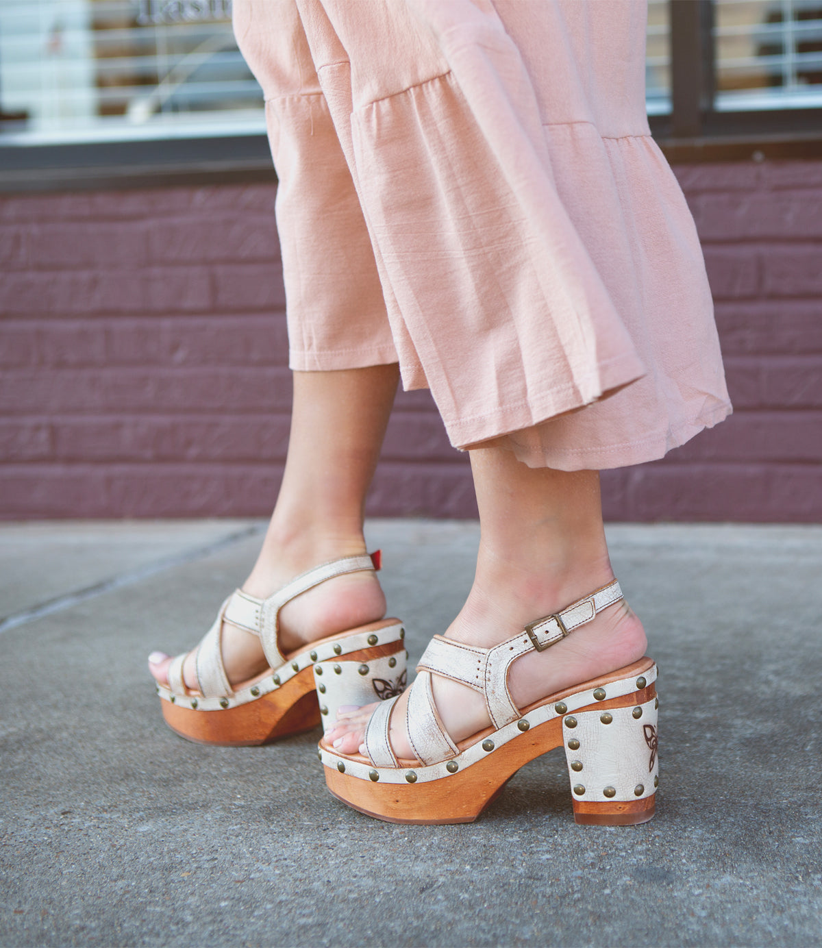 Wearing the Bed|Stü Mediation beige heeled slingback sandals with cushioned insoles and wooden soles, a person in light pink wide-leg pants stands on a sidewalk near a brick wall.