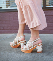 Wearing the Bed|Stü Mediation beige heeled slingback sandals with cushioned insoles and wooden soles, a person in light pink wide-leg pants stands on a sidewalk near a brick wall.